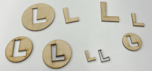 Laser cut pieces of the Luigi “L” logo of varying sizes, some as outlines of the “L”, some as just the “L”, and some as circles with the “L” cut out of the middle laid out on a white background. 